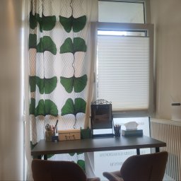 Leśne Słówka - Gabinet terapeutyczny z biurkiem, krzesłami i zasłonami w liście miłorzębu. Widoczne akcesoria biurowe i okno z żaluzjami. Neutralna kolorystyka ścian.