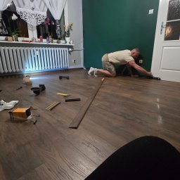 Miro BOSS_BUD - Montaż listew przypodłogowych w Barlinku. Fachowiec na kolanach przy drzwiach, używa poziomicy. Na podłodze narzędzia i akcesoria montażowe. Nowe panele podłogowe.