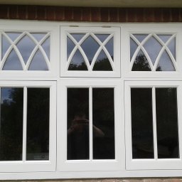 Timber Windows - Białe okno z ozdobnymi szprosami w kształcie rombów, zamontowane w murze z czerwonej cegły. W szybach odbija się osoba robiąca zdjęcie.