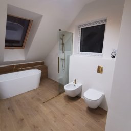 EDBUD.COMPANI - Jasna łazienka na poddaszu ze skośnym sufitem. Biała wanna, kabina prysznicowa ze złotymi detalami, bidet i toaleta. Podłoga wyłożona płytkami imitującymi drewno.