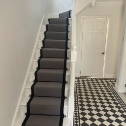 sppserwis - Białe schody z szarym dywanem i czarnymi akcentami. W tle podłoga w czarno-białą szachownicę i białe drzwi. Minimalistyczny design.