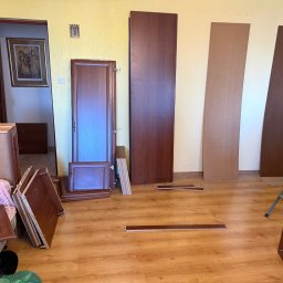 Multiserwis - Demontaż mebli w pokoju z drewnianą podłogą. Widoczne elementy szafy, drzwi, drabina i osoba w tle. Przygotowanie do transportu mebli.