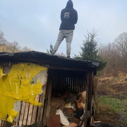 Multiserwis - Mężczyzna w bluzie z kapturem stoi na dachu zrujnowanej szopy, wypełnionej śmieciami. W tle drzewa i pochmurne niebo. Widoczny fragment żółtej plandeki.