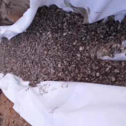 Wykop wypełniony kamieniami, otulony białą geowłókniną, z widoczną rurą drenarską i studzienką kontrolną w tle.