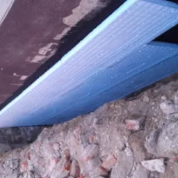 Prace przy ocieplaniu fundamentów w Częstochowie: płyty izolacyjne z widoczną siatką na fragmentach fundamentu, ziemia i gruz w wykopie.