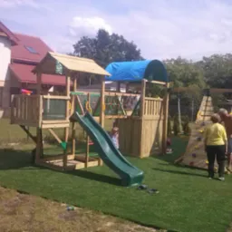 Drewniany plac zabaw z wieżą, zjeżdżalnią, ścianką wspinaczkową i huśtawką, ustawiony na sztucznej trawie w Trzcielu, z rodziną w tle. Widoczny dom z czerwonym dachem.