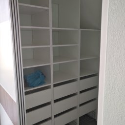 LUXURY FURNITURE - Biała szafa wnękowa z półkami i szufladami, widok z wnętrza. Na jednej z półek leży niebieska torba. Widoczne metalowe kosze na dole szafy.