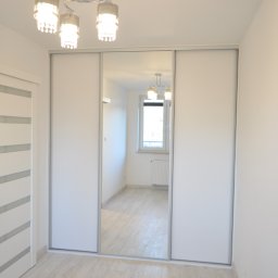 LUXURY FURNITURE - Biała szafa przesuwna z lustrem w minimalistycznym wnętrzu. Widoczne oświetlenie sufitowe i fragment drzwi z przeszkleniami. Odbicie pokoju w lustrze.
