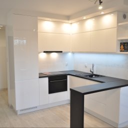 LUXURY FURNITURE - Biała, nowoczesna kuchnia z czarnym blatem i barkiem. Górne szafki podświetlone, na półce dekoracja. Widoczna drewniana podłoga i ciemne drzwi.
