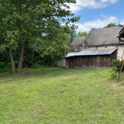 Patryk Juszczyk - Świeżo skoszony trawnik z widocznymi ścinkami trawy, rozciągający się przed starym, drewnianym budynkiem gospodarczym z blaszanym dachem, obok kamiennego domu z dachówką, otoczony zielonymi...