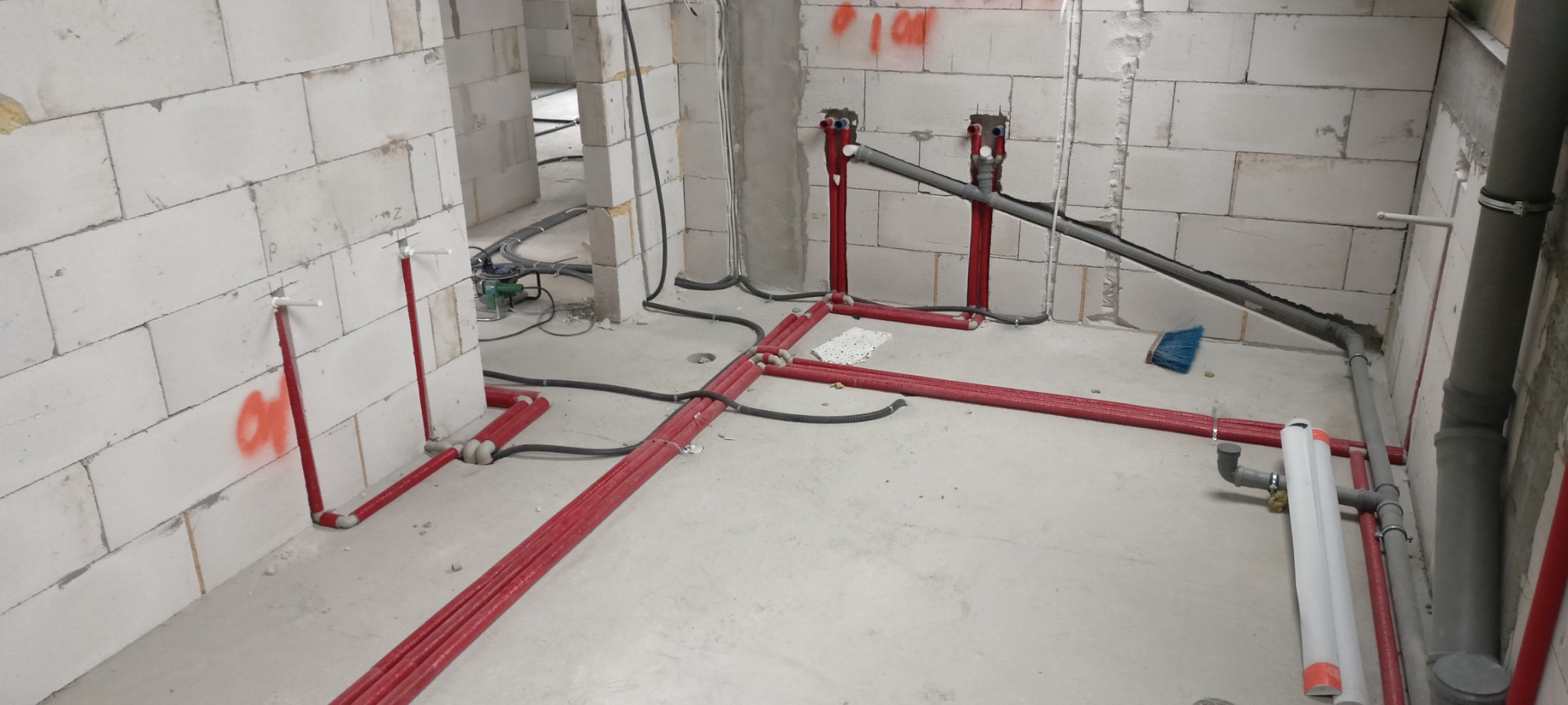 Instalacja hydrauliczna i grzewcza w surowym stanie: czerwone rury na podłodze i przy ścianach z białego bloczka, szare rury kanalizacyjne, widoczne kable i narzędzia.