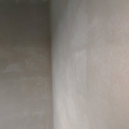 Pat Tech - Ściana po nałożeniu tynku maszynowego, widoczne nierówności i faktura. Ujęcie z dołu, fragment sufitu z elementami konstrukcyjnymi.