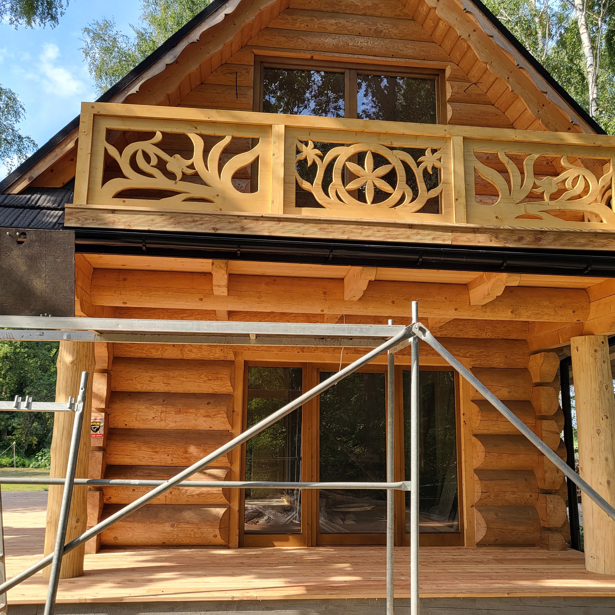 Surowa drewniana konstrukcja domu z bali z dekoracyjną balustradą balkonu, widoczne rusztowanie. Detal wykończenia, naturalne drewno, częściowo zadaszony dach.