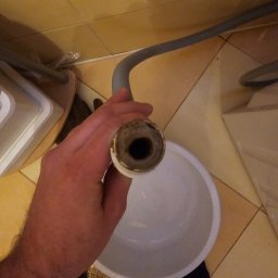 Flush czyszczenie kanalizacji - Zbliżenie na dłoń trzymającą rurę z grubym, ciemnym osadem wewnątrz, nad białym wiadrem na tle beżowych płytek.