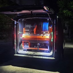 Flush czyszczenie kanalizacji - Tył ciemnego vana z otwartą klapą, ukazujący specjalistyczne, pomarańczowo-czarne urządzenie do czyszczenia rur z dwoma szpulami węży, oświetlone w nocy.
