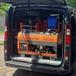 Flush czyszczenie kanalizacji - Tył otwartego czarnego busa z zamontowaną pomarańczowo-szarą maszyną do czyszczenia instalacji, wyposażoną w dwie szpule z wężami (czarny i niebieski) oraz panel sterowania. Bus stoi na bruku.