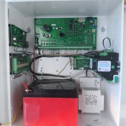 Bartosz Lewicki - Otwarta, biała skrzynka z elektroniką alarmową: płyty główne, kable, akumulator i transformator. Widoczne komponenty marki Satel i Pulsar.