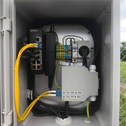 Bartosz Lewicki - Wnętrze skrzynki telekomunikacyjnej z elementami instalacji światłowodowej i elektrycznej, Poznań. Widoczne kable, złączki i urządzenia.