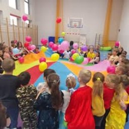Turbiki Katarzyna Filarek-Kocurek - Dzieci bawią się kolorową chustą animacyjną w sali gimnastycznej, unosząc balony. Widoczne kosze do koszykówki i okna. Radosna atmosfera zabawy grupowej.