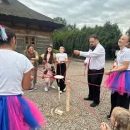 Turbiki animacje fotomagnesy na żywo - Grupa dzieci i dorosłych gra w grę z czerwoną liną na zewnątrz, w tle drewniany budynek i pochmurne niebo. Zabawa integracyjna z animatorem.