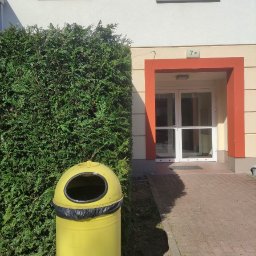 Jerzy Dmytriv - Żółty kosz na śmieci z symbolem segregacji stoi przed równo przyciętym żywopłotem z tui, obok wejścia do budynku z numerem 7B w słoneczny dzień.