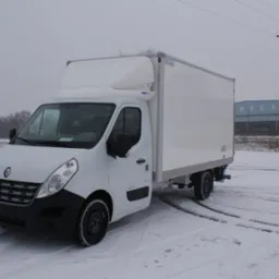 Biały samochód dostawczy Renault Master z zabudową kontenerową, zaparkowany na śniegu, z widocznymi śladami opon. Na drugim planie budynek przemysłowy i drzewo bez liści.