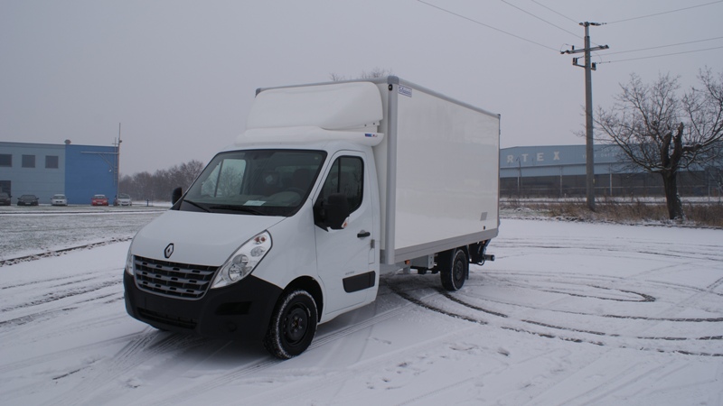 Biały samochód dostawczy Renault Master z zabudową kontenerową, zaparkowany na śniegu, z widocznymi śladami opon. Na drugim planie budynek przemysłowy i drzewo bez liści.