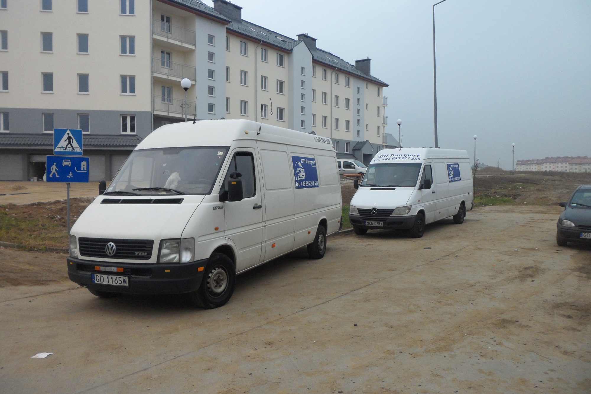 Dwa białe busy firmy transportowej zaparkowane na placu budowy przed blokiem mieszkalnym w pochmurny dzień. Na bokach busów widoczne logo i numer telefonu.