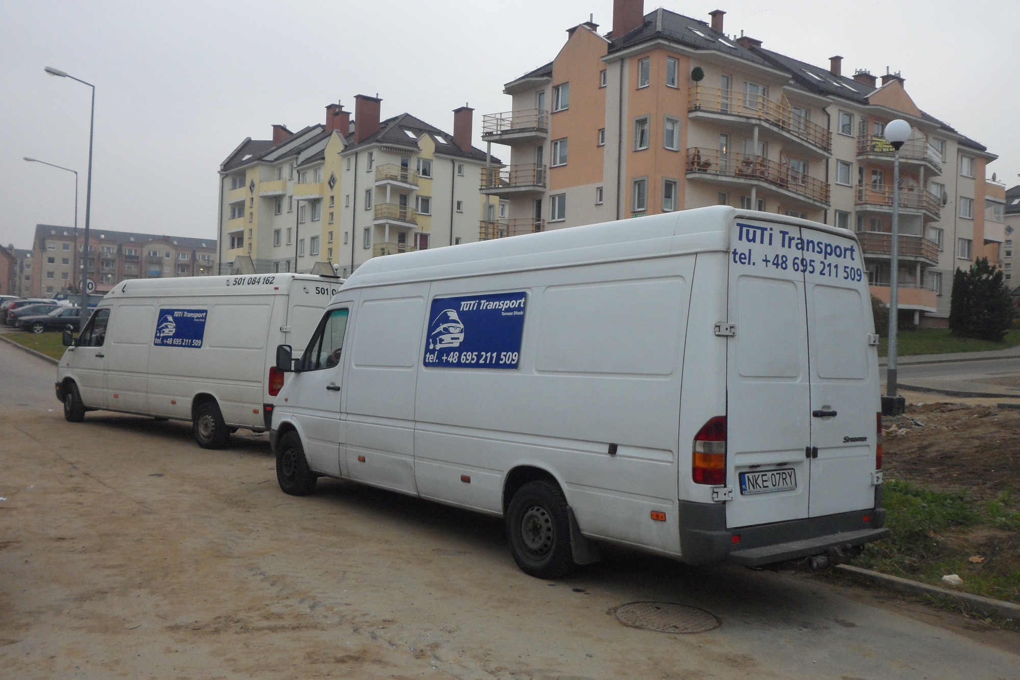 Dwa białe busy firmy transportowej z logo i numerem telefonu zaparkowane na betonowym placu przed budynkami mieszkalnymi w pochmurny dzień. Na tylnej części busa widoczna tablica rejestracyjna.