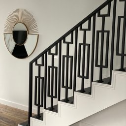 MetalCraft - Nowoczesna, czarna balustrada metalowa z geometrycznym wzorem na białych schodach. Na ścianie dekoracyjne lustro w stylu art deco.