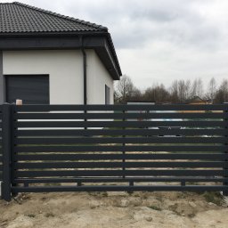 MetalCraft - Nowoczesne, poziome ogrodzenie metalowe w kolorze grafitowym przed domem z szarą dachówką. W tle widoczny samochód i drzewa bez liści.