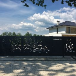 MetalCraft - Nowoczesna, czarna brama wjazdowa z ażurowym wzorem drzewa i adresem 'Wiśniowa 21', na tle domu z jasną elewacją i błękitnego nieba z chmurami.