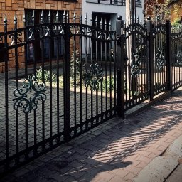 MetalCraft - Czarna, metalowa brama z dekoracyjnymi elementami na tle ceglanego budynku i kostki brukowej. Widoczne cienie rzucane przez bramę.