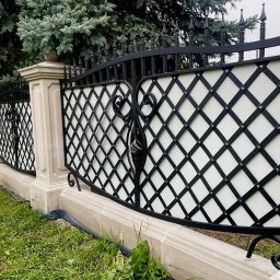 MetalCraft - Eleganckie ogrodzenie metalowe z białym wypełnieniem w romby, osadzone na kamiennej podmurówce, otoczone zielenią. Widok z perspektywy trawnika.