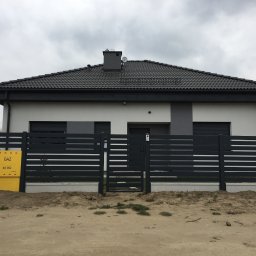 MetalCraft - Nowoczesne, poziome ogrodzenie metalowe w kolorze antracytowym przed domem z szarą elewacją i żółtą skrzynką gazową. Minimalistyczny design, pochmurne niebo.
