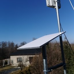 Piotr Kondraciuk Zakład Usług Komputerowych Kaspio - Płaska, biała antena satelitarna i szare urządzenie sieciowe zamontowane na metalowym maszcie na tle błękitnego nieba, z domem jednorodzinnym i drzewami w oddali.