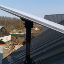 Piotr Kondraciuk Zakład Usług Komputerowych Kaspio - Biała, prostokątna antena satelitarna zamontowana na czarnym maszcie do ciemnoszarego, blaszanego dachu. W tle wiejski krajobraz z drzewami i zabudowaniami.