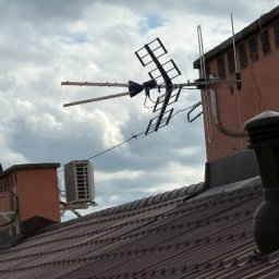 Piotr Kondraciuk Zakład Usług Komputerowych Kaspio - Duża antena telewizyjna i mniejsza antena na dachu pokrytym brązową dachówką, obok zewnętrzna jednostka klimatyzacji, na tle zachmurzonego nieba.