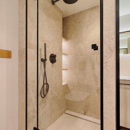 ARCHITEKSTURA Malwina Kroll architekt - Elegancka kabina prysznicowa walk-in z czarnymi okuciami i beżowymi płytkami imitującymi kamień. Minimalistyczny design, oświetlenie punktowe, półki wnękowe.