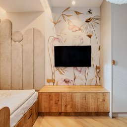 ARCHITEKSTURA Malwina Kroll architekt - Pokój dziecięcy z tapetą w kwiaty i motyle, beżowym łóżkiem z tapicerowanym zagłówkiem oraz drewnianą szafką RTV. Subtelne oświetlenie i jasna kolorystyka.