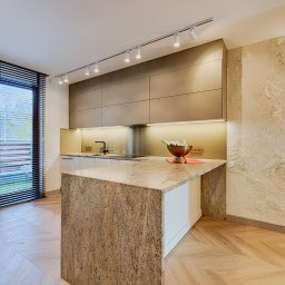 ARCHITEKSTURA Malwina Kroll architekt - Nowoczesna kuchnia z wyspą z kamienia, oświetleniem punktowym i parkietem w jodełkę. Ściana z dekoracyjnym tynkiem. Okno z żaluzjami w tle.