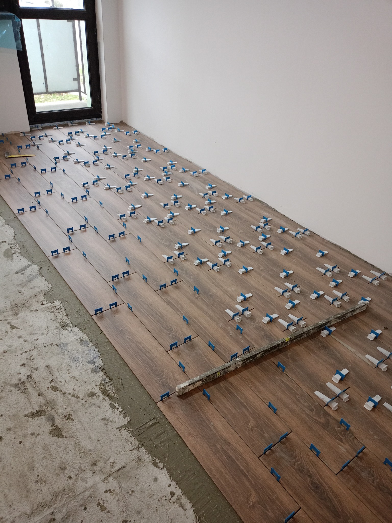 Układanie drewnopodobnych płytek podłogowych z użyciem poziomicy i klipsów poziomujących w jasnym pomieszczeniu z oknem.