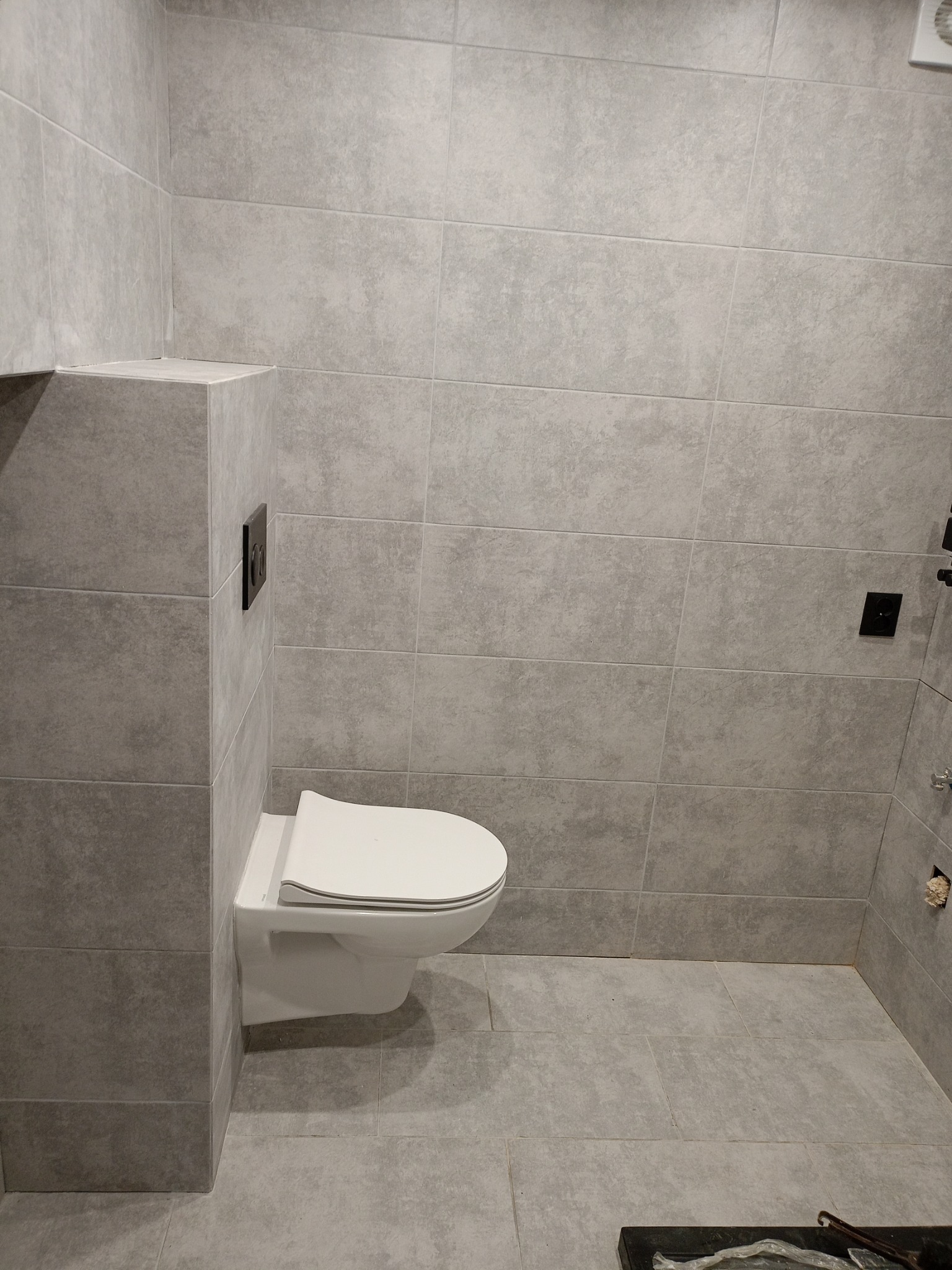 Nowoczesna toaleta podwieszana w szarej, wykafelkowanej łazience z czarnymi przyciskami spłukiwania i widoczną wentylacją.