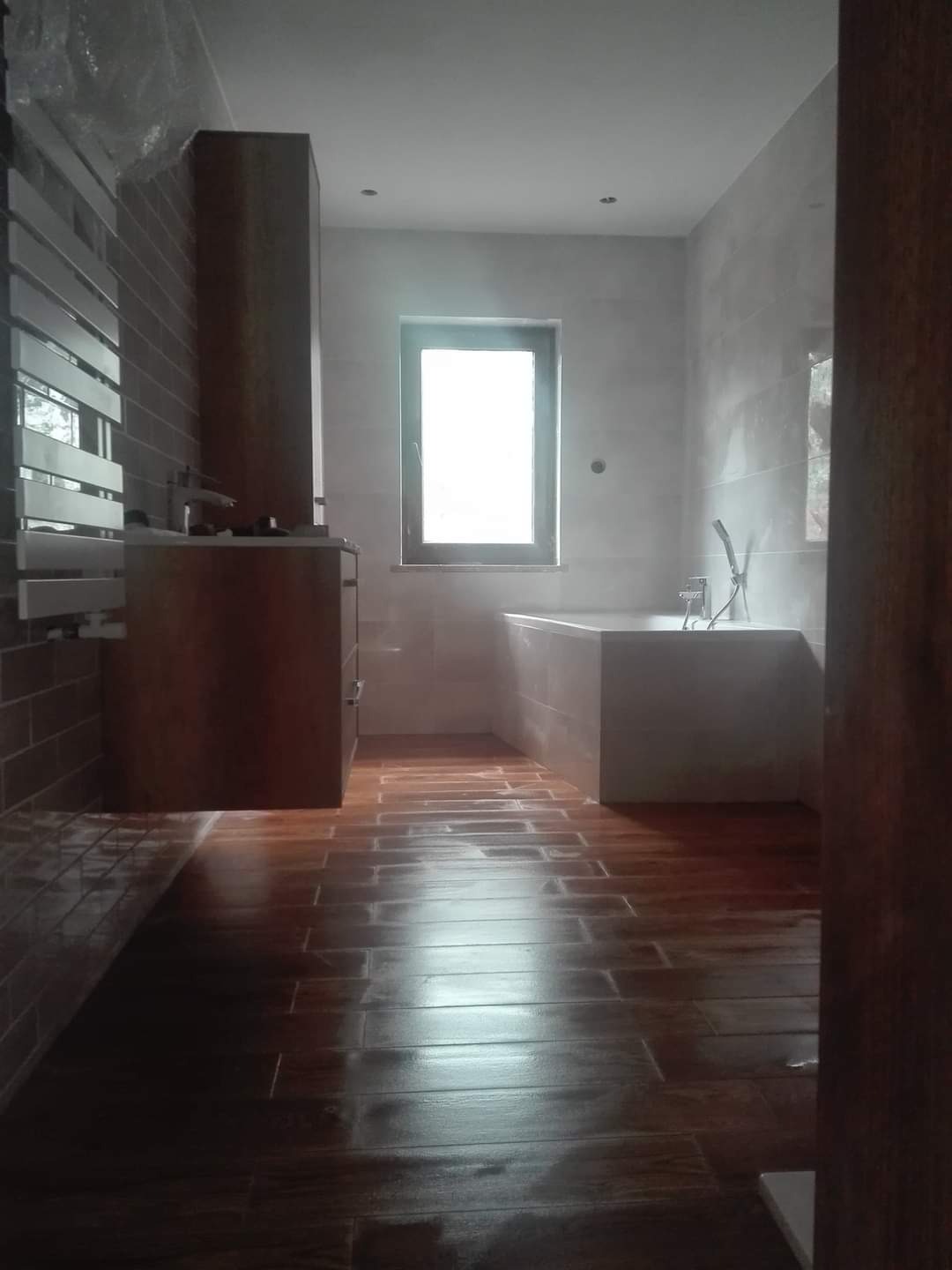 Nowoczesna łazienka z brązowymi płytkami podłogowymi imitującymi drewno, jasnymi ścianami i zabudowaną wanną przy oknie, widoczna szafka z umywalką.