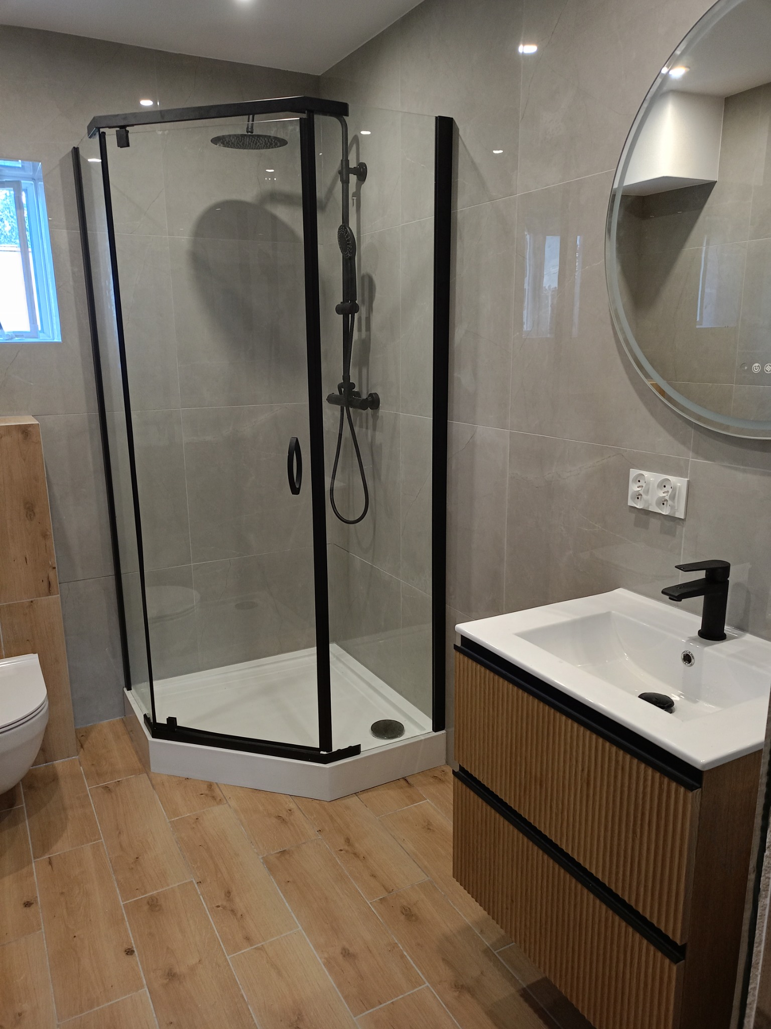 Nowoczesna łazienka z kabiną prysznicową narożną z czarnymi profilami, umywalką nablatową na szafce z ryflowanym frontem i okrągłym lustrem z podświetleniem LED.