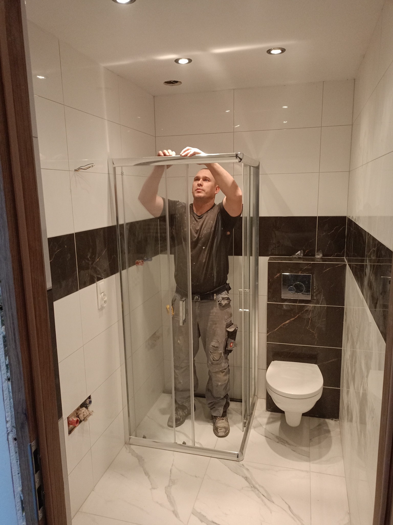 Hydraulik instaluje kabinę prysznicową z chromowanymi profilami w łazience z białymi i brązowymi płytkami, widoczny fragment instalacji wodnej w ścianie.