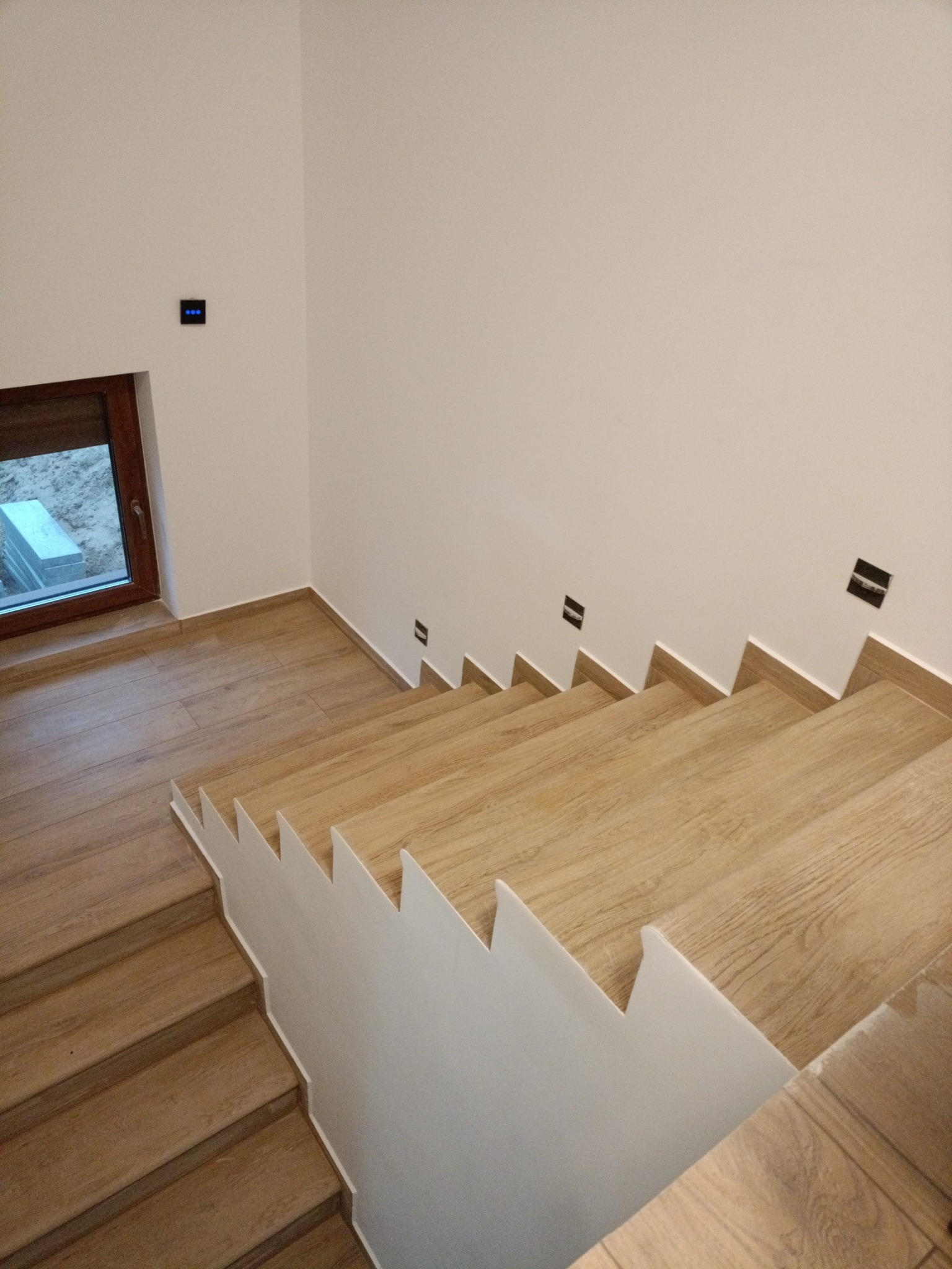 Schody wewnętrzne wykończone panelami imitującymi drewno, biała ściana z czarnymi gniazdkami, widok z góry.