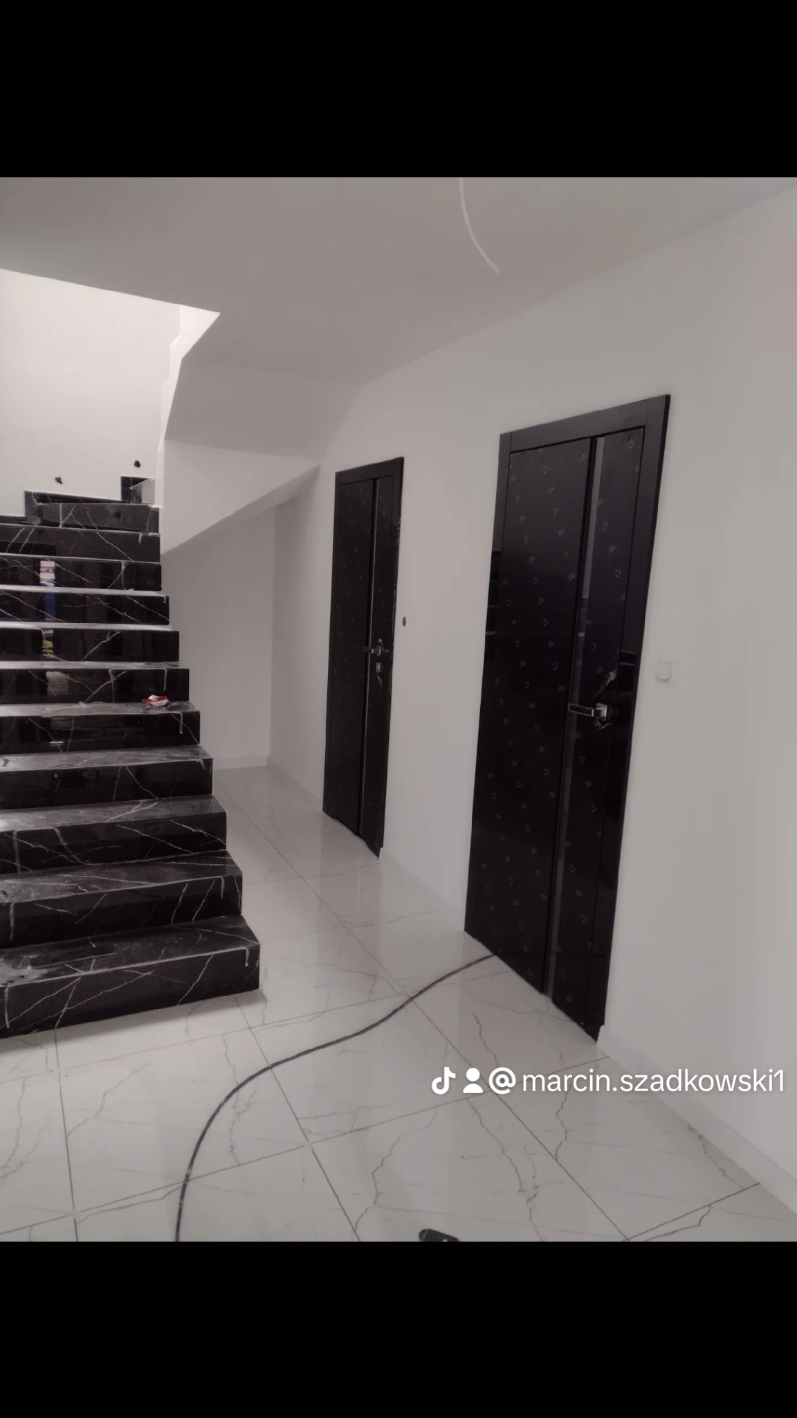 Nowoczesne wnętrze z czarnymi schodami marmurowymi i dwoma czarnymi drzwiami z delikatnym wzorem na białym tle ścian i podłogi z białych płytek imitujących marmur.