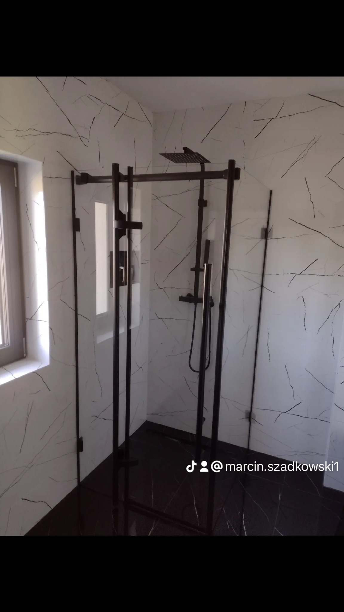 Nowoczesna kabina prysznicowa z czarnymi profilami na tle ściany z imitacją marmuru, czarna podłoga, widok z boku, okno z lewej strony.