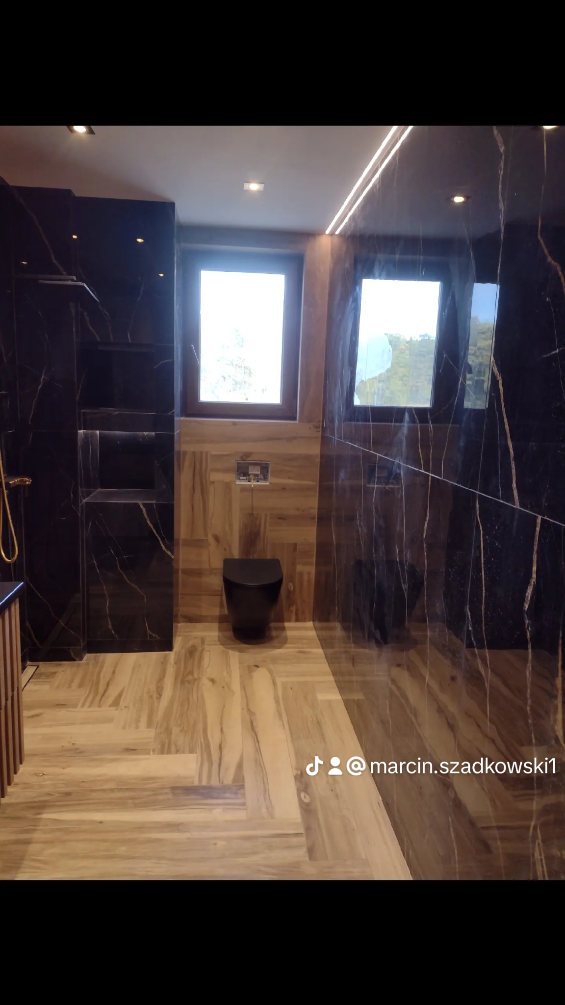 Nowoczesna łazienka z czarnymi marmurowymi ścianami i drewnopodobną podłogą ułożoną w jodełkę, podświetlane półki w ścianie, czarna wisząca toaleta, dwa okna w naturalnym drewnie.